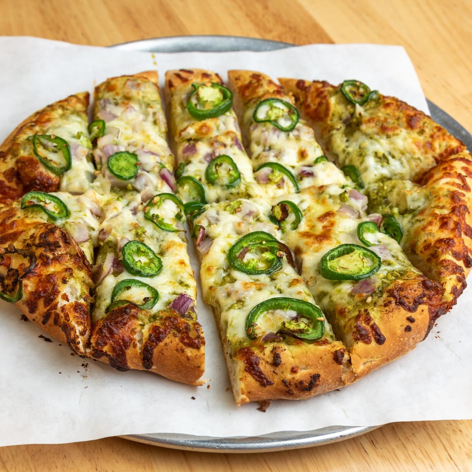 A sliced pesto pizza with melted cheese, red onions, and green jalapeño slices on a metal tray.