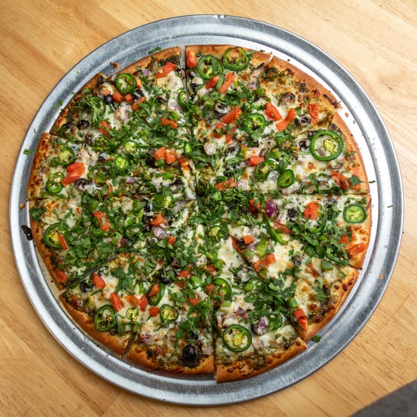 A whole pizza on a metal tray, topped with cheese, jalapenos, tomatoes, olives, and cilantro.